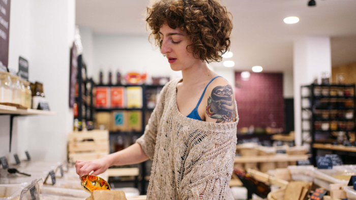 Eine Frau mit lockigem Haar und einem Tattoo auf ihrem Oberarm füllt in einem Unverpackt-Laden Nudeln in eine Papiertüte. Sie trägt einen beigen, gestrickten Pullover und steht vor einer Theke mit verschiedenen Behältern und Regalen voller Produkte. Die Umgebung wirkt freundlich und ordentlich, mit natürlicher Beleuchtung und einer gemütlichen Atmosphäre.