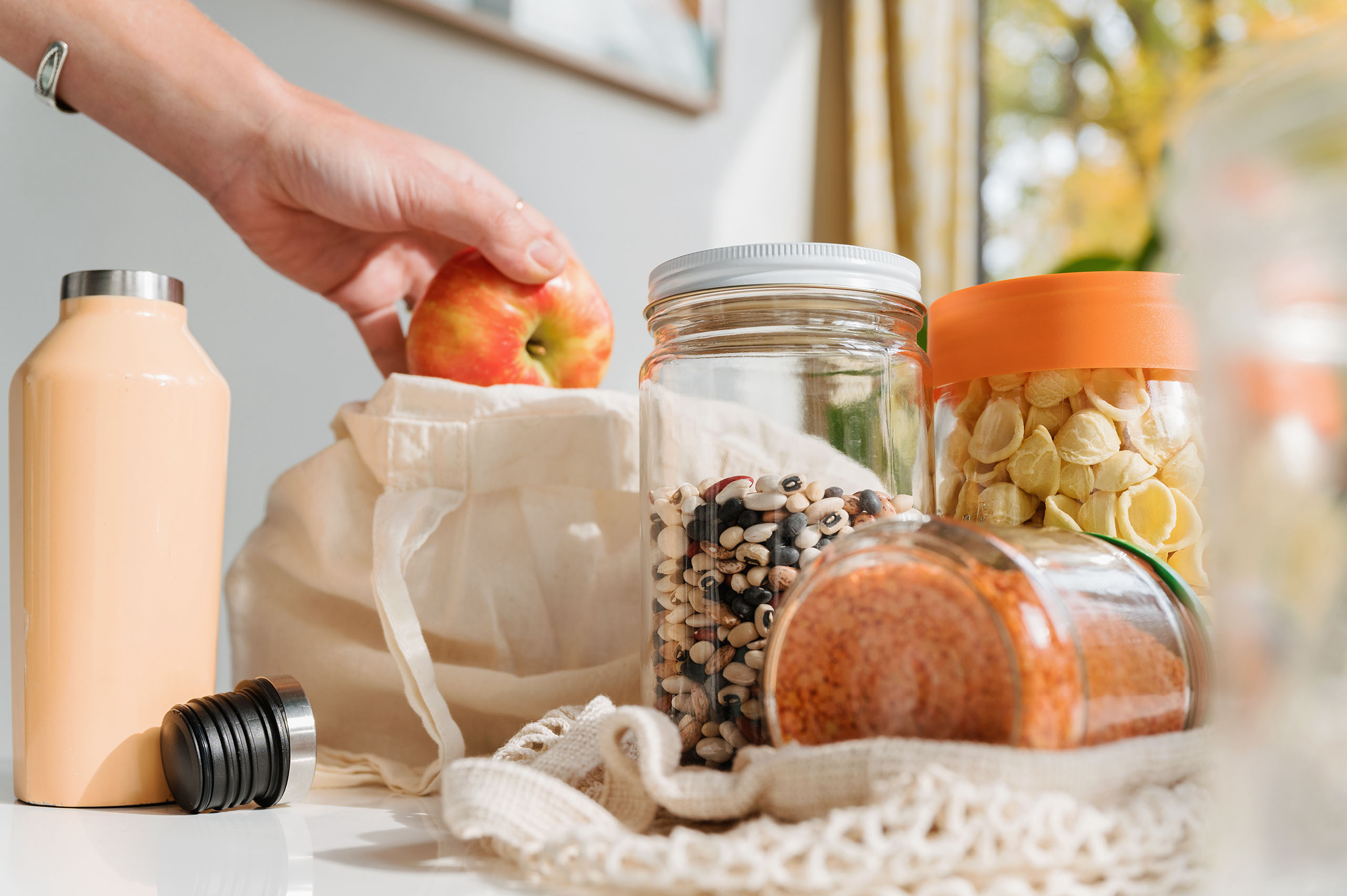 Zu sehen sind Gläser mit Bohnen, Linsen und Nudeln, eine Aluminiumtrinkflasche und eine Hand die einen Apfel aus einer Stofftasche nimmt