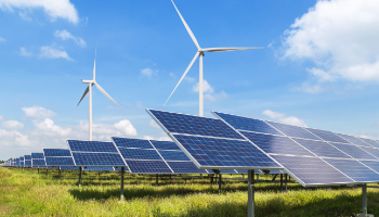 Eine große Solaranlage steht auf einer grünen Wiese unter einem blauen Himmel mit ver-einzelten weißen Wolken. Im Hintergrund stehen Windräder.