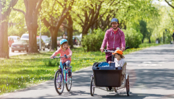 Ein Vater fährt mit einem Lastenrad auf einem Radweg, ein Kind fährt neben ihm auf einem Fahrrad. Ein zweites Kind sitzt im Lastenrad. Es ist sonnig, im Hintergrund sind geparkte Autos und Bäume zu sehen.