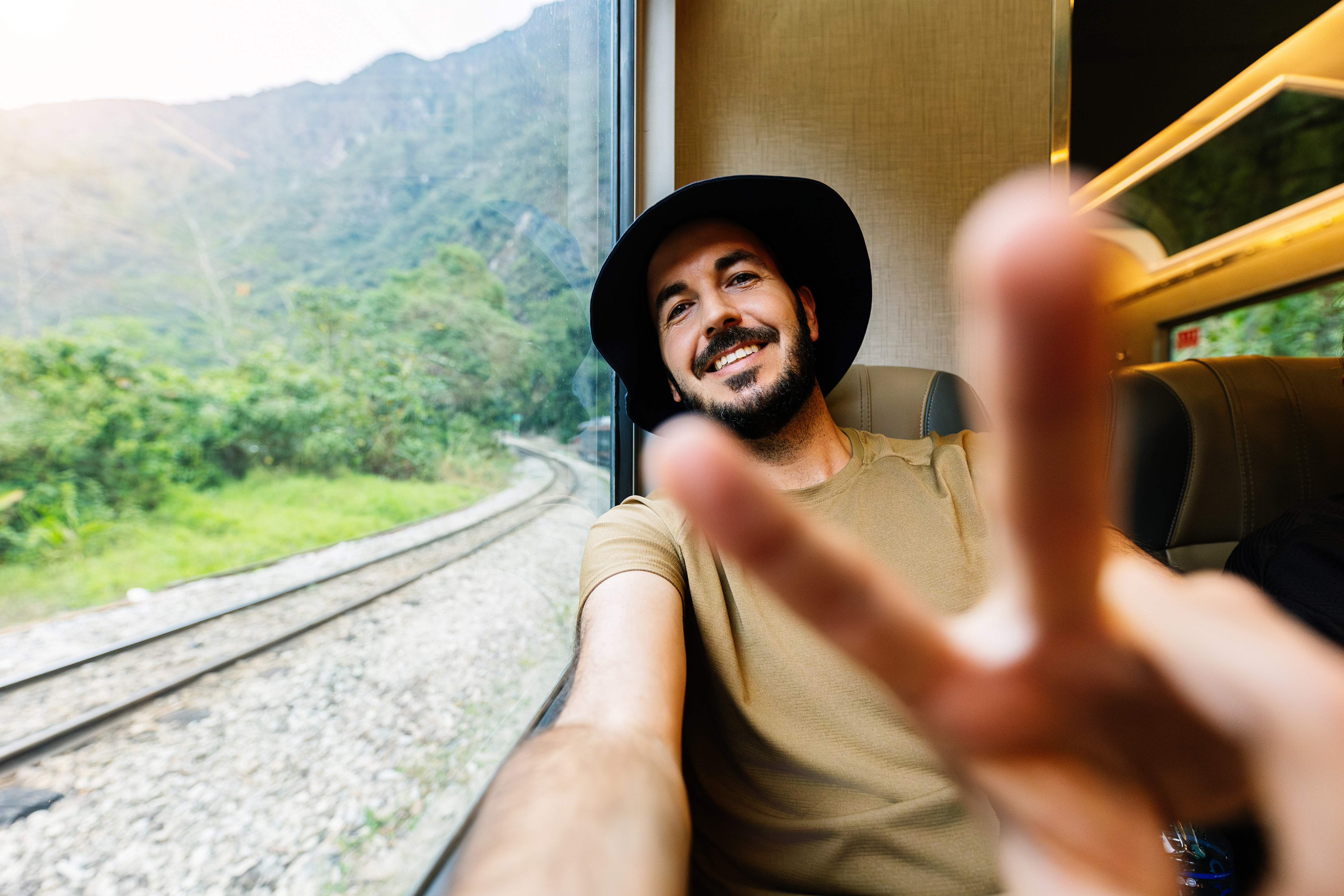 Ein lächelnder Mann mit schwarzem Hut sitzt in einem Zug am Fenster und macht ein Selfie. Im Vordergrund ist unscharf seine Hand mit einem Peace-Zeichen zu sehen, während draußen grüne Berge und Bahngleise vorbeiziehen.
