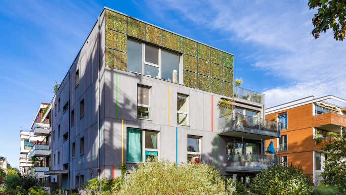 Ein modernes Wohngebäude mit grauer Fassade, bunten vertikalen Streifen, einer Mooswand im oberen Stockwerk und Balkonen, umgeben von Pflanzen.