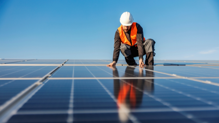 Ein Arbeiter mit weißem Schutzhelm und orangefarbener Warnweste kniet auf einer großen Fläche von Solarpaneelen unter blauem Himmel.