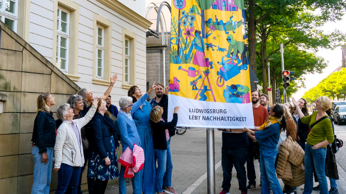 Beteiligte Akteurinnen und Akteure zeigen auf die „Nachhaltigkeitsflagge“ vor dem Rathaus und freuen sich.