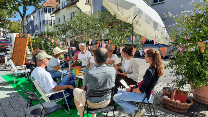 Menschen unterschiedlichen Alters sitzen gemeinsam an einem Campingtisch auf einem Parkplatz in der Sonne unter einem Sonnenschirm.