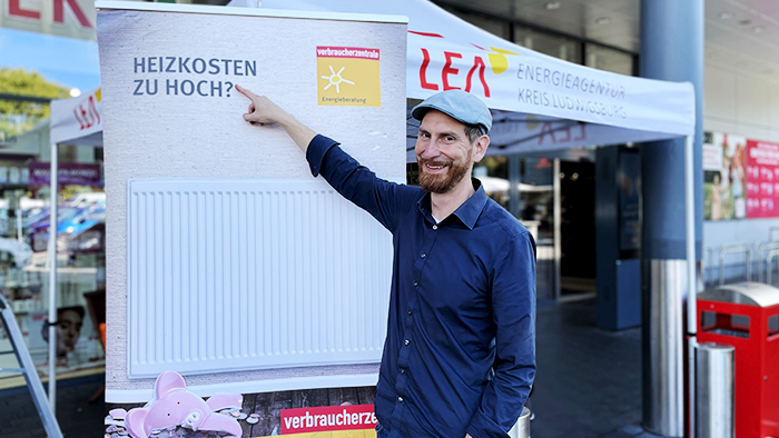 Mann mit Baskenmütze, der lächelnd auf ein Banner zeigt, auf dem „Heizkosten zu hoch?" steht.