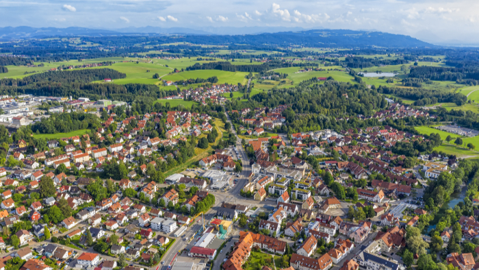 Luftaufnahme einer kleinen Stadt inmitten einer grünen Landschaft mit weitläufigen Feldern und Wäldern im Hintergrund.