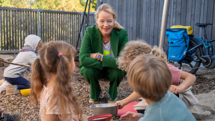 Umweltministerin Thekla Walker geht in die Hocke und spricht mit Kindern, die draußen im Garten des Waldorfkindergartens spielen.
