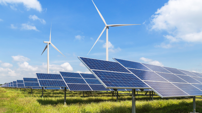 Eine große Solaranlage steht auf einer grünen Wiese unter einem blauen Himmel mit vereinzelten weißen Wolken. Im Hintergrund stehen Windräder.